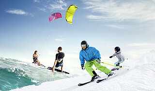 Im Vordergund sind zwei Personen am Snowboarden imSchnee. im Hintergrund sind zwei Personen, die auf dem Wasser wakeboarden   