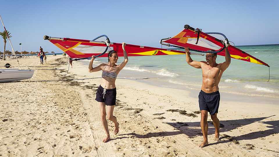 Eine Frau und ein Mann tragen ihr Surfsegel vom Meer auf den Strand.