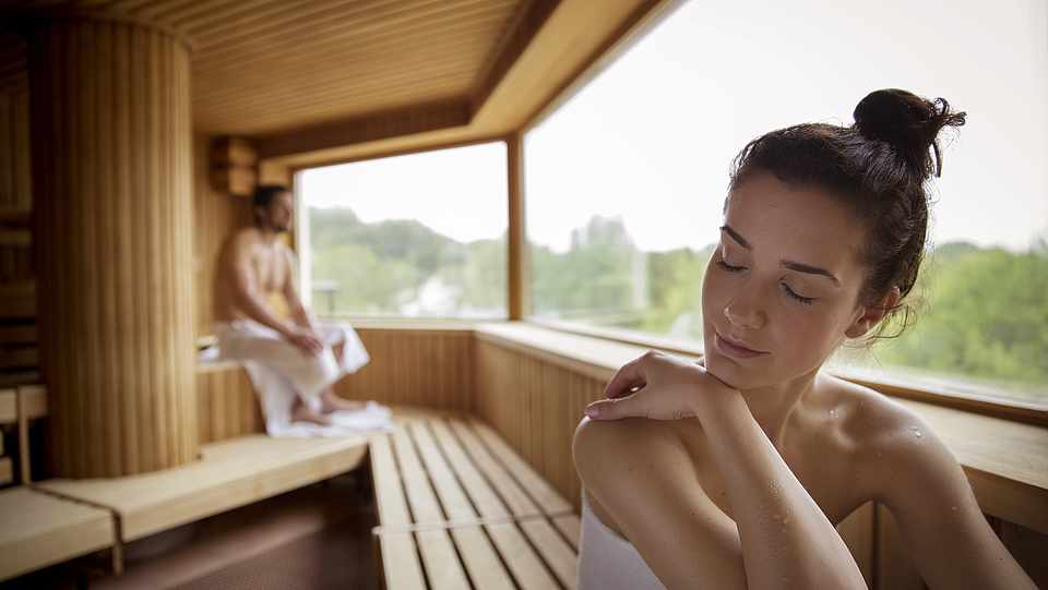 Eine Frau sitzt im Bildvordergrund auf einer Saunabank. Im Hintergrund sitzt ebenfalls ein Mann. In der Sauna gibt es ein Panorama-Fenster.