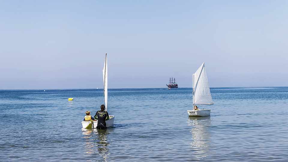 In einem kleinen Boot mit Segel sitzen ein Junge und ein Mann. etwas weiter rechts ist ein weiteres kleines Segelboot. Zwischen den beiden Segeln etwas weiter links sieht man ein großes Holzschiff in der Ferne.
