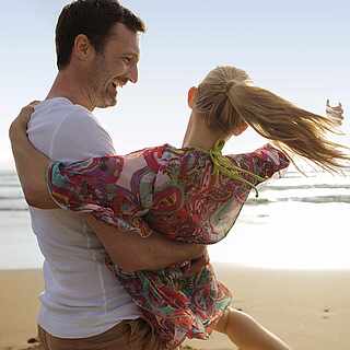 Am Strand sind ein Mann und eine Frau, wobei der Mann die Frau auf den Armen hält und sie sich zusammen im Kreis drehen