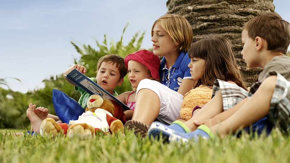 Mehrere Kinder sitzen zusammen und kriegen von der Betreuerin ein Buch vorgelesen