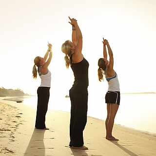 Drei Frauen führen eine Yoga-Figur aus. Sie stehen am Strand nahe des Wassers. Links sieht man Wald.