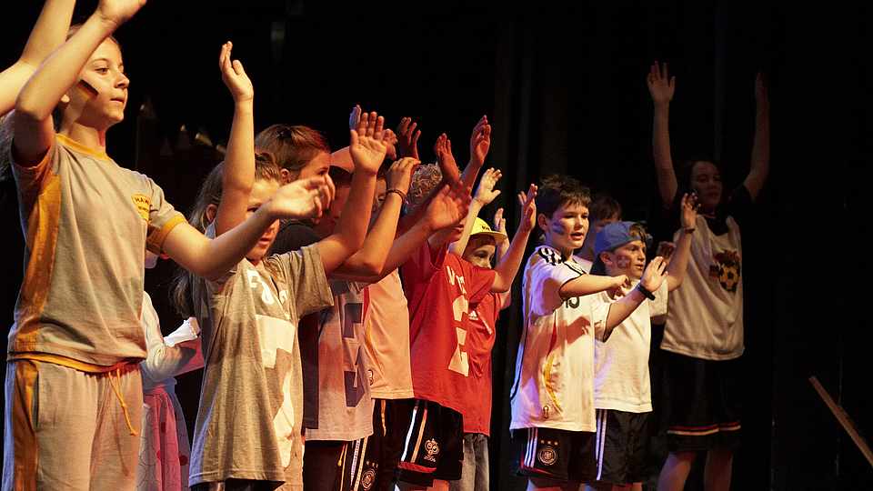 Kinder stehen nebeneinander auf der Bühne und heben ihre Hände hoch
